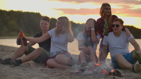 The-young-people-are-sitting-in-around-bonfire-on-the-river-coast.-They-are-laughing-and-singing-songs-with-beer-at-sunset-in-summer-evening.
